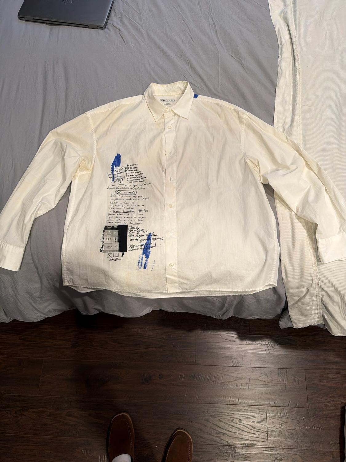 White shirt with handwritten text and blue stains on a grey bed. Wooden floor and tips of shoes in a relaxed setting.