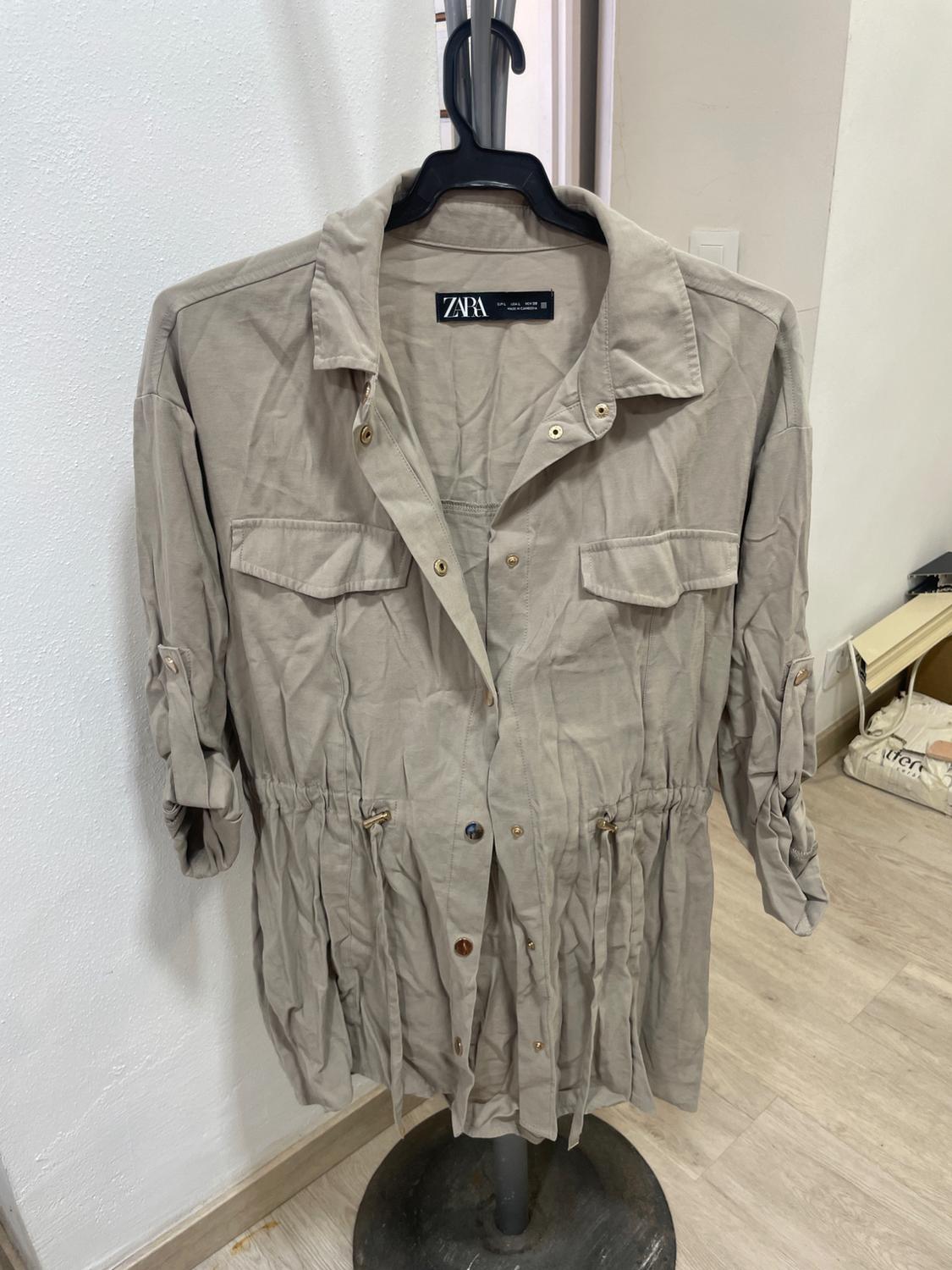 Beige, long-sleeved jacket with front pockets and an adjustable drawstring waist, hanging on a black hanger.