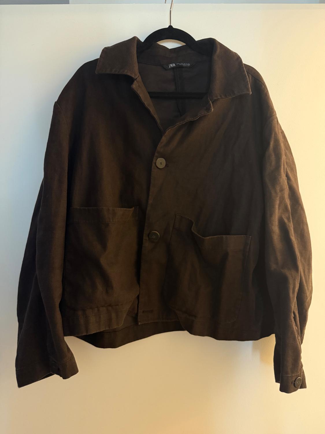 Brown, thick fabric jacket hanging on a black hanger. It has two large pockets and front buttons.