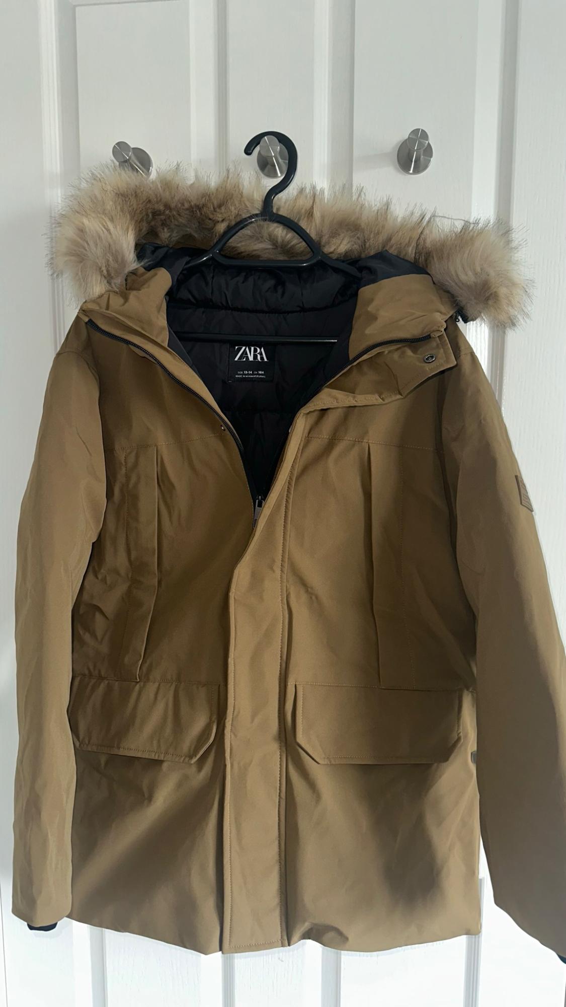 Brown parka coat with a faux fur hood hanging on a hanger over a white door. Label with the text ZARA.