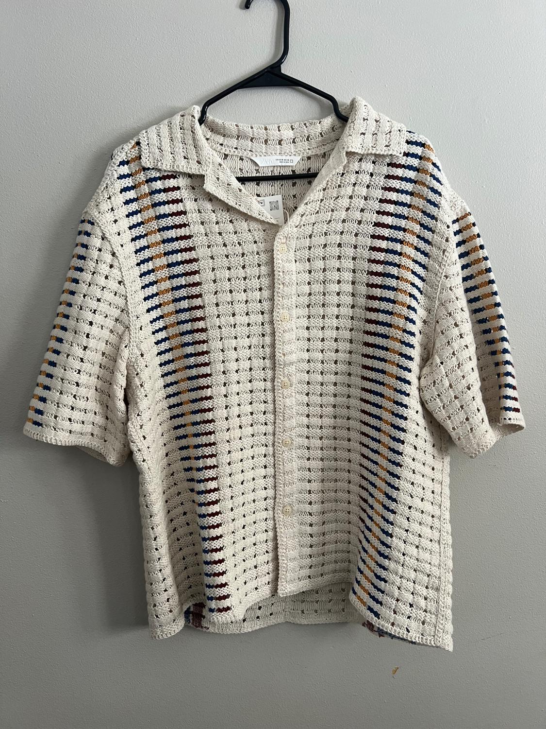 Beige knit shirt with vertical blue, brown, and mustard stripes, hanging on a black hanger against a gray wall.