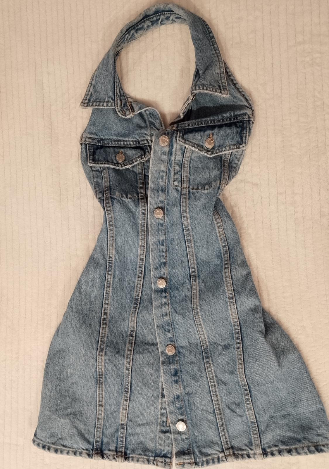 Light blue denim dress with a halter neck, front buttons and pockets, laid out on a light fabric background.