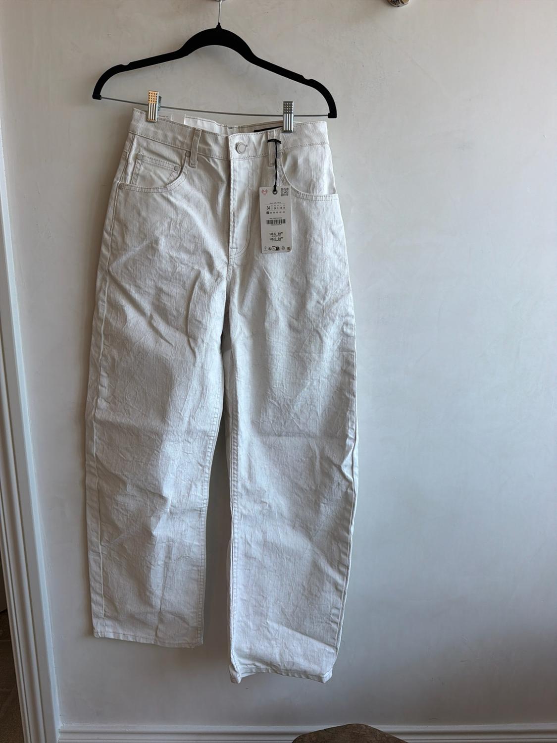 White straight-leg jeans hanging from a black hanger with metal clips against a white wall.