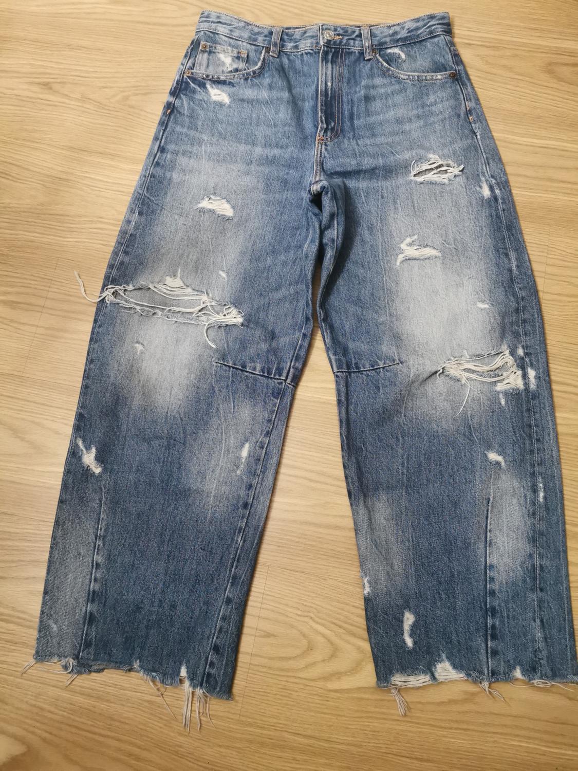 Casual style blue denim jeans with rips, a distressed effect, and a frayed hem, laid out on a light wooden floor.