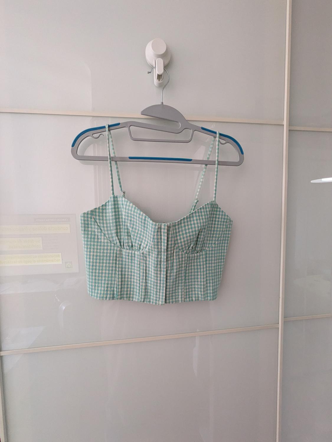Green and white checked bustier-style crop top with thin straps, hanging on a hanger over a white door.