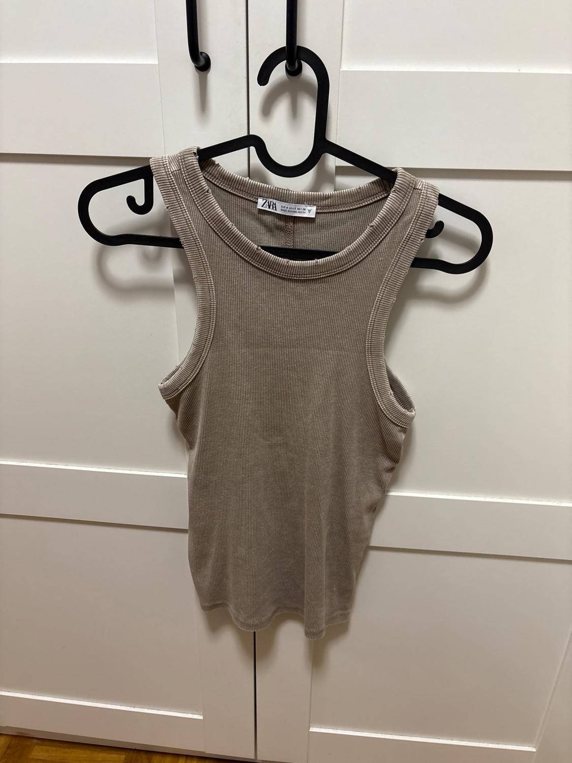 Beige ribbed vest top with ZARA text on the label, hanging on a black hanger over a white wardrobe. Casual style.