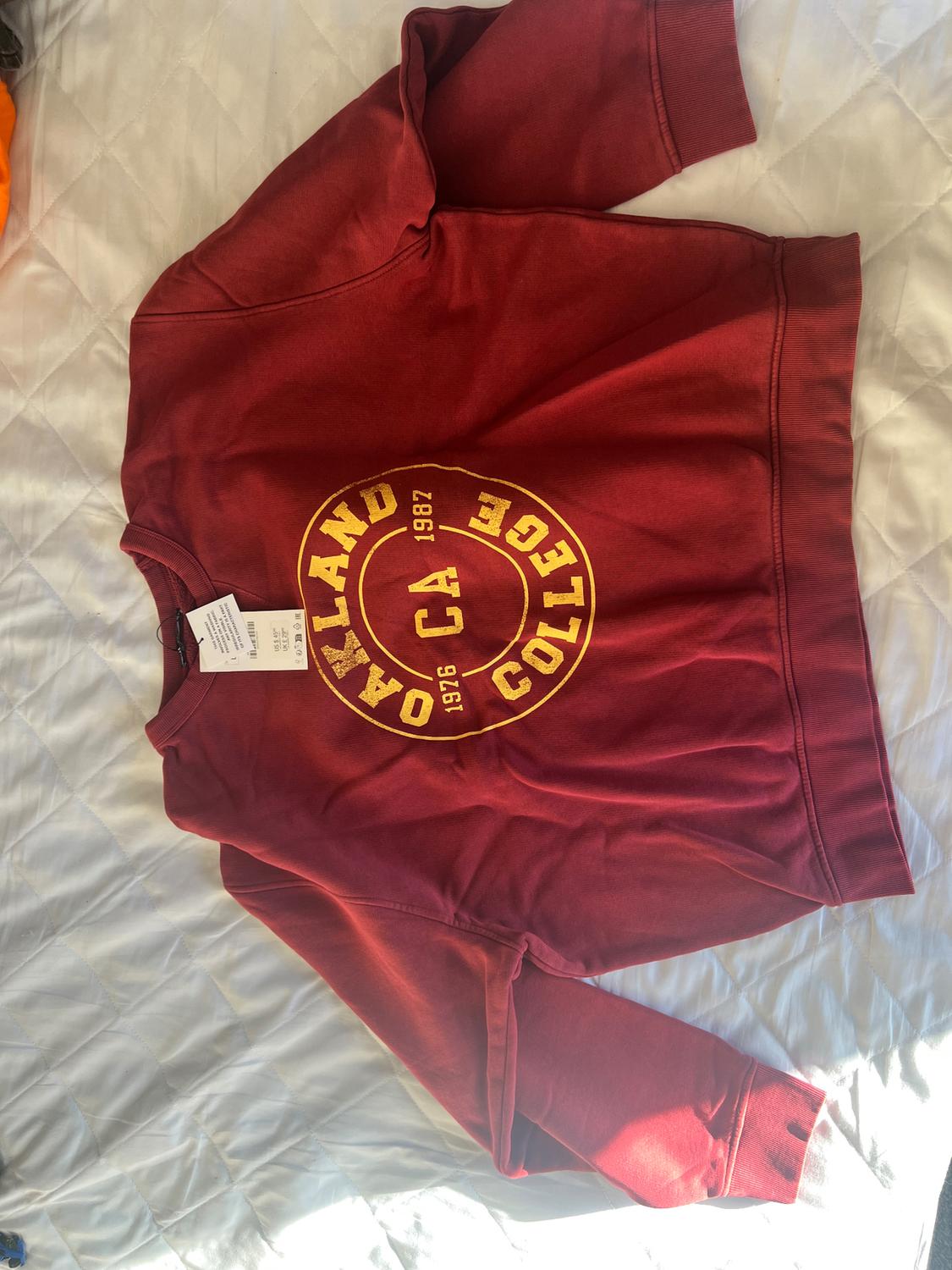 Red sweatshirt with a yellow "OAKLAND COLLEGE 1976 1987" print, on a white bed with a visible label.