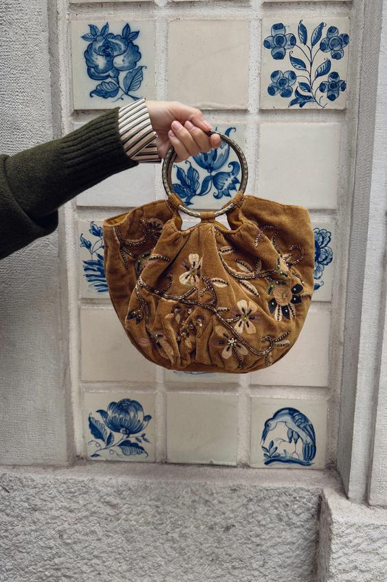 Detail hnd kabelky s kvtinovou vivkou a kovovmi kruhovmi uchy. Viditeln zelen rukv svetru.