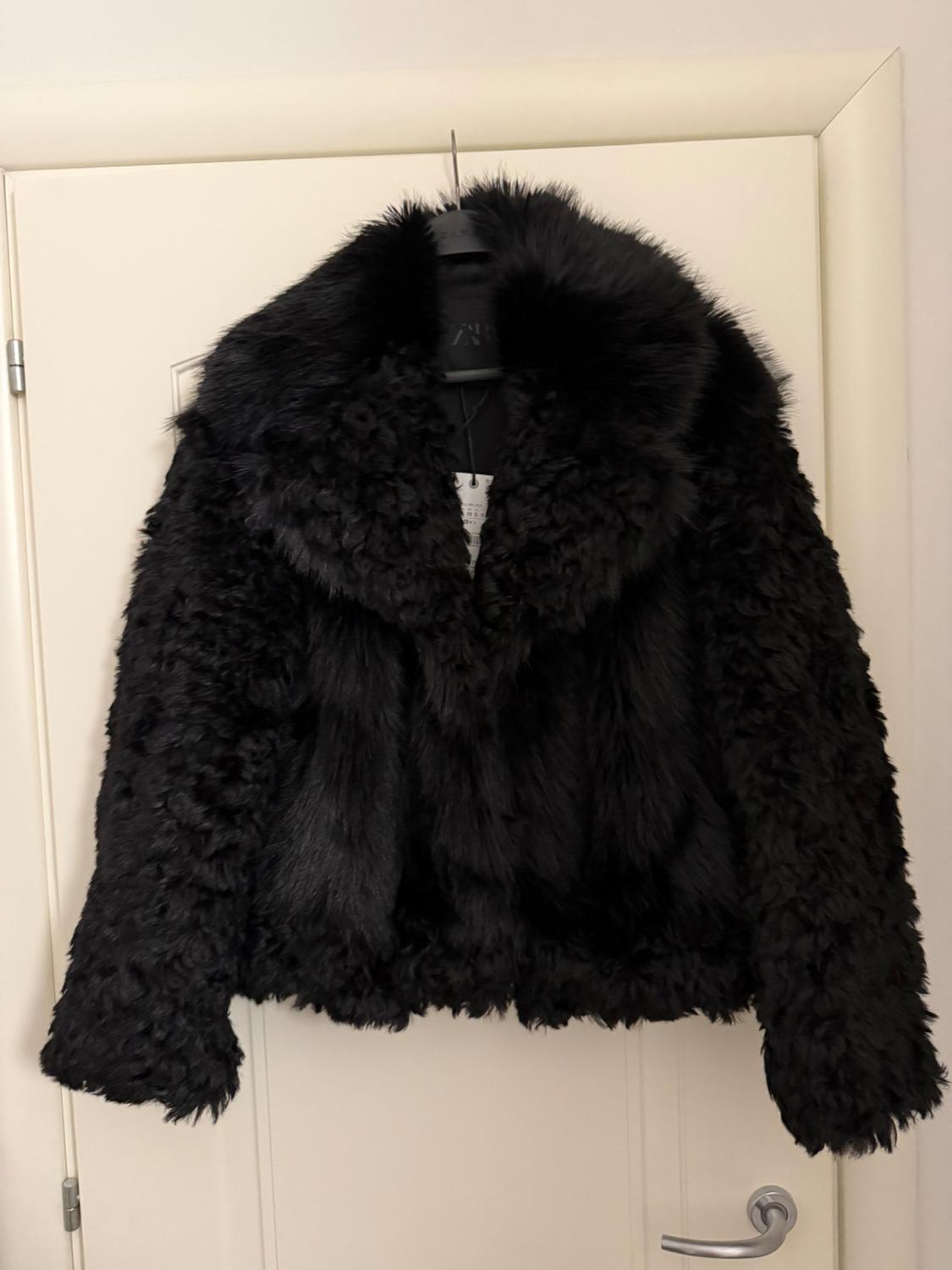 Black faux leather jacket hanging on a white door. Fluffy and voluminous design with a wide collar.
