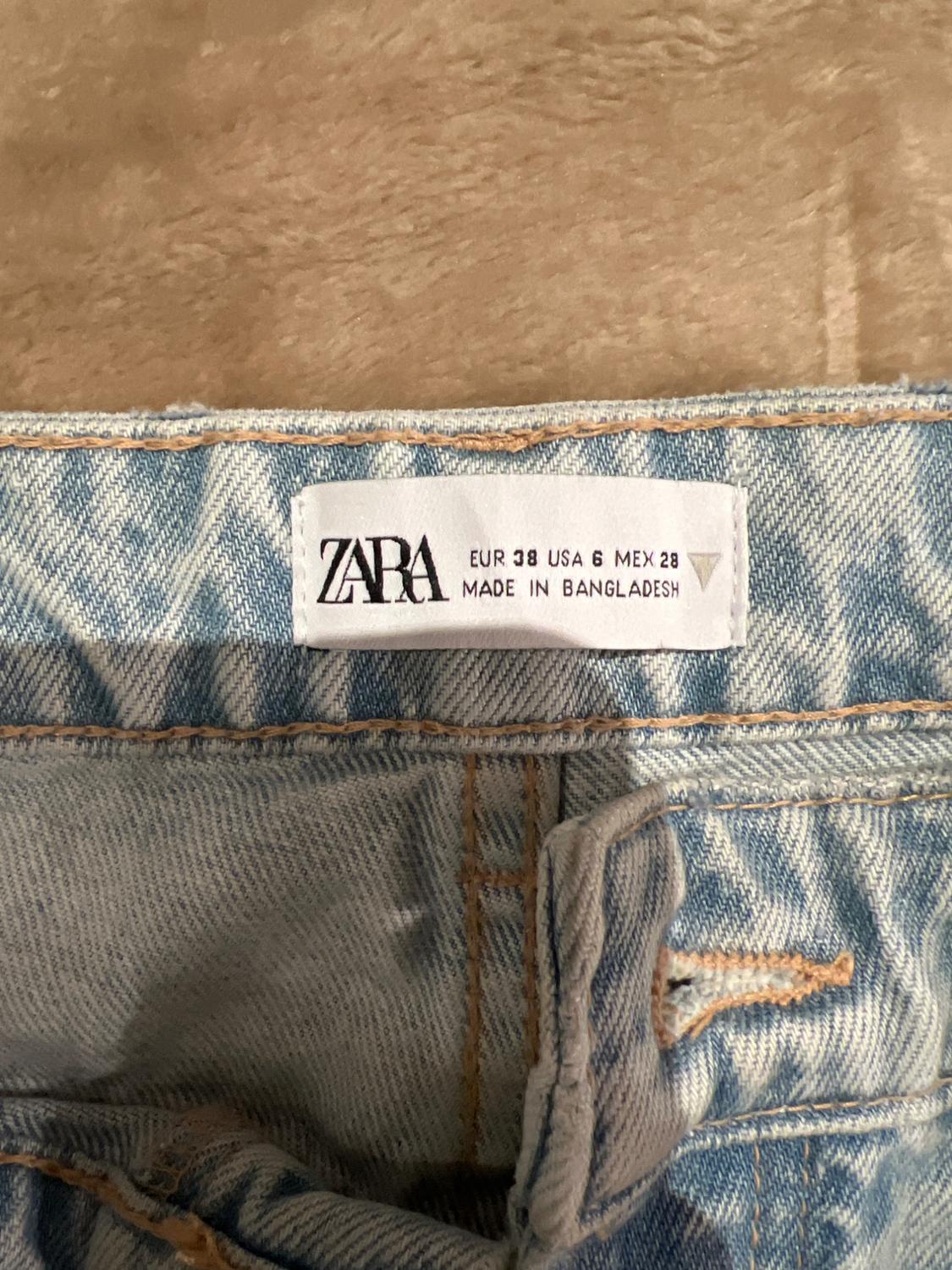 Blue jeans label with brown stitching. Visible text: "ZARA EUR 38 USA 6 MEX 28 MADE IN BANGLADESH". Beige background.