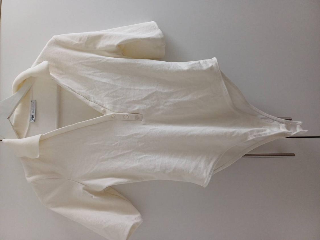 White, short-sleeved bodysuit with a collar and buttons, hanging on a hanger against a white background. Label visible on the collar.