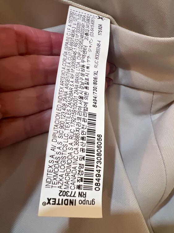 A hand holds an Inditex clothing tag with text in several languages on a beige fabric. At the end, a barcode can be seen.
