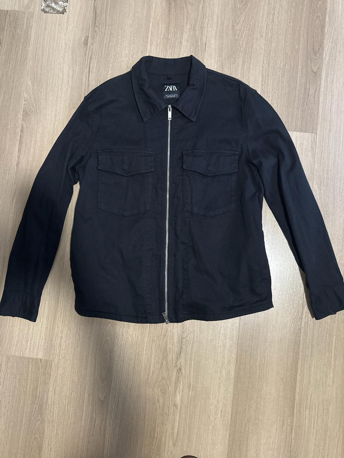 Black jacket with a front zip and chest pockets, laid out on a wooden floor. The inner label reads ZARA.