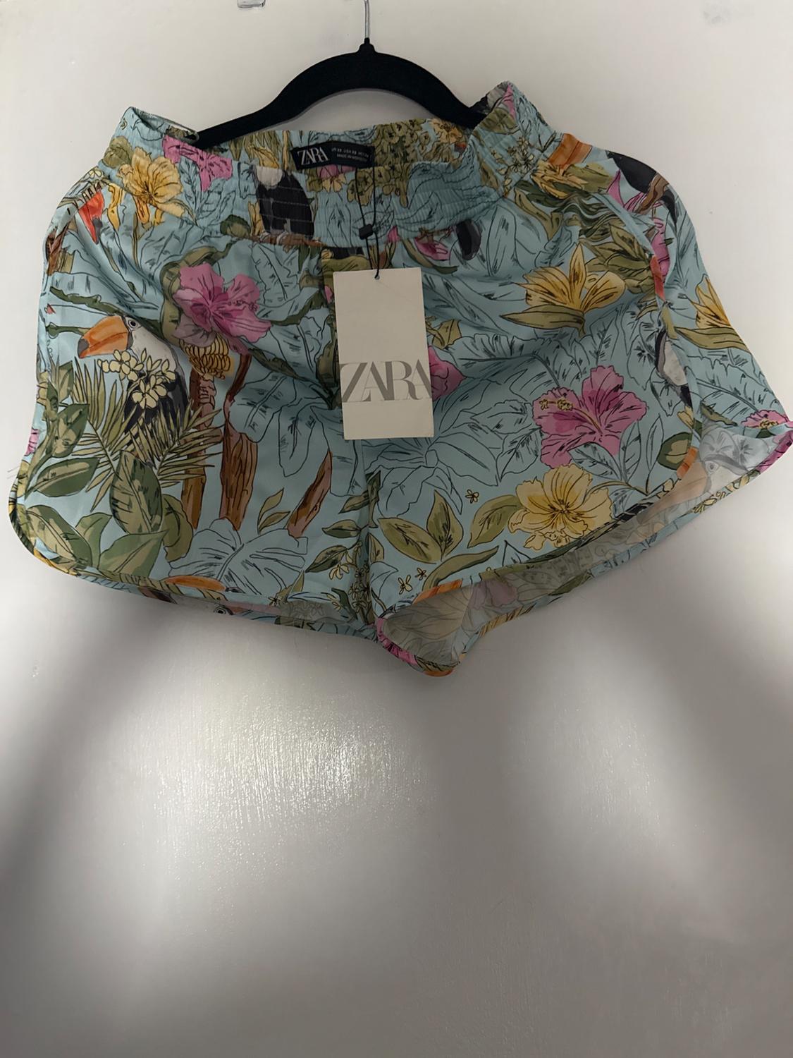 Shorts with a floral print in pastel tones, hanging on a black hanger. Visible tag with the text "ZARA". White background.