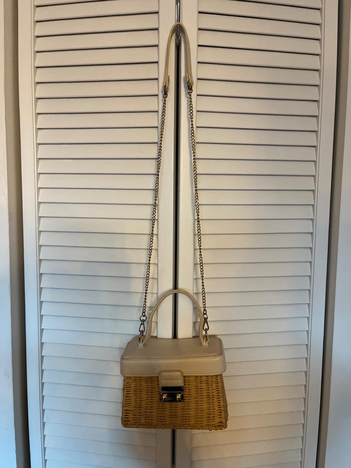 Wicker and beige leather handbag hanging on a white shutter door, with a gold chain and metal clasp.