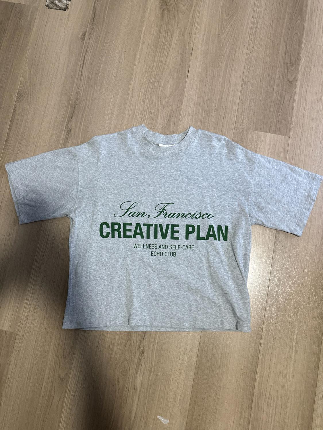 Grey short-sleeved T-shirt on a wooden floor. Text: San Francisco CREATIVE PLAN WELLNESS AND SELF-CARE ECHO CLUB.