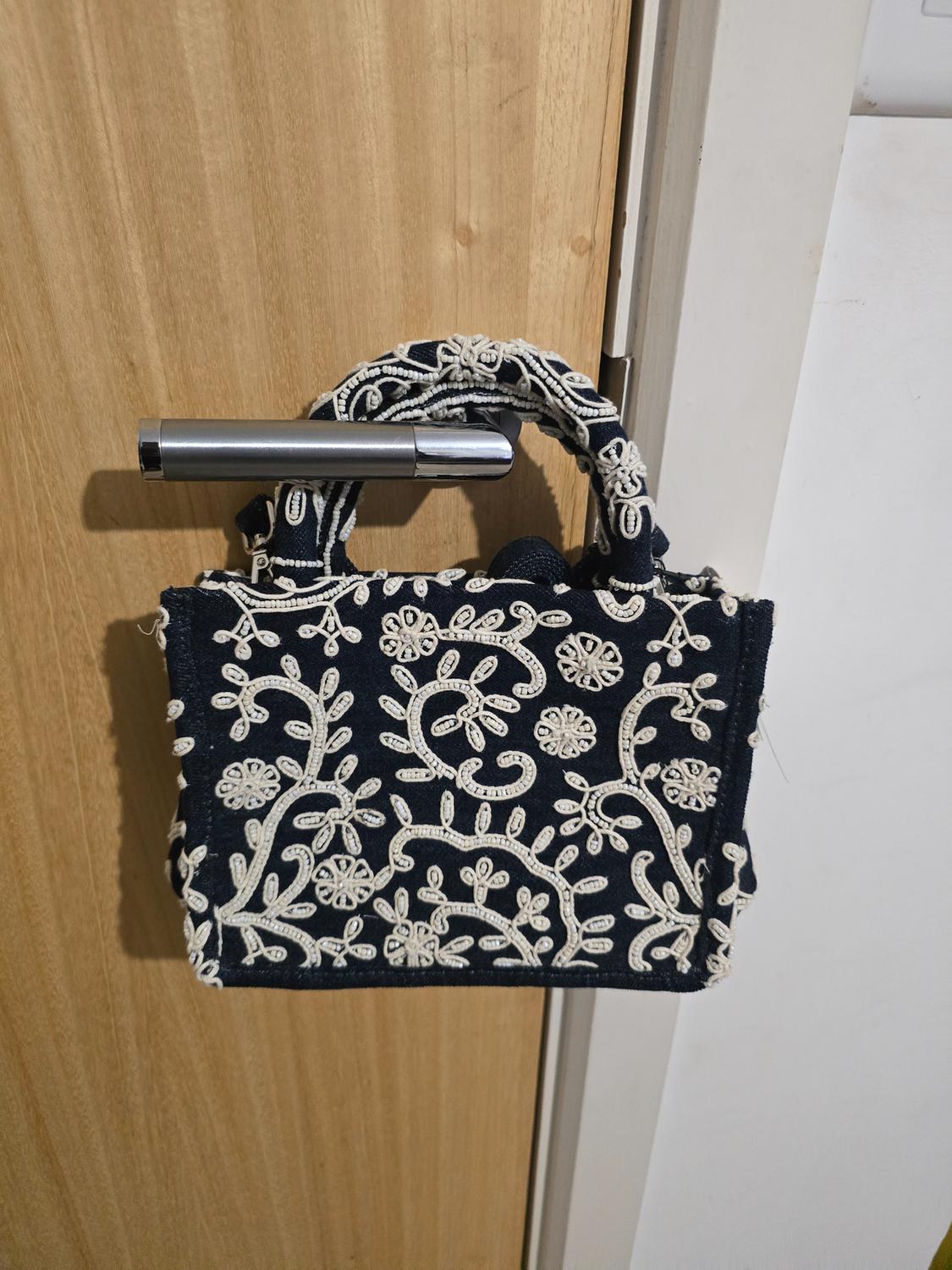 Small blue handbag with white floral embroidery hanging from a silver door handle on a light wooden door.