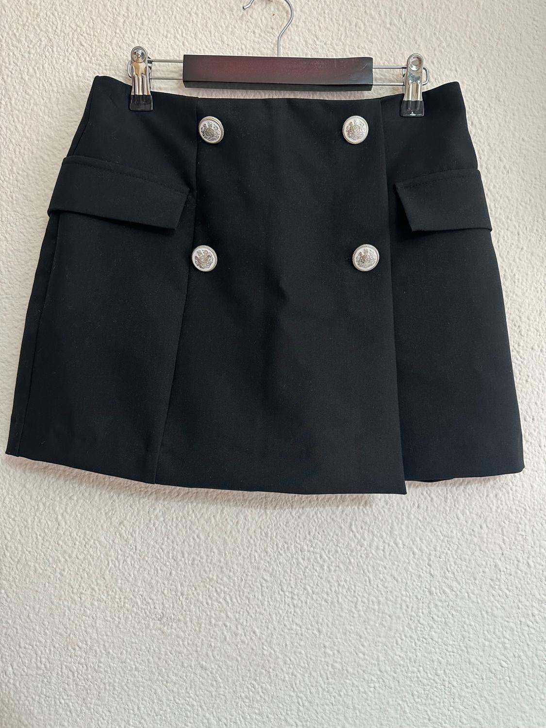 Short black skirt with silver buttons and front pockets, hanging on a hanger against a white wall.