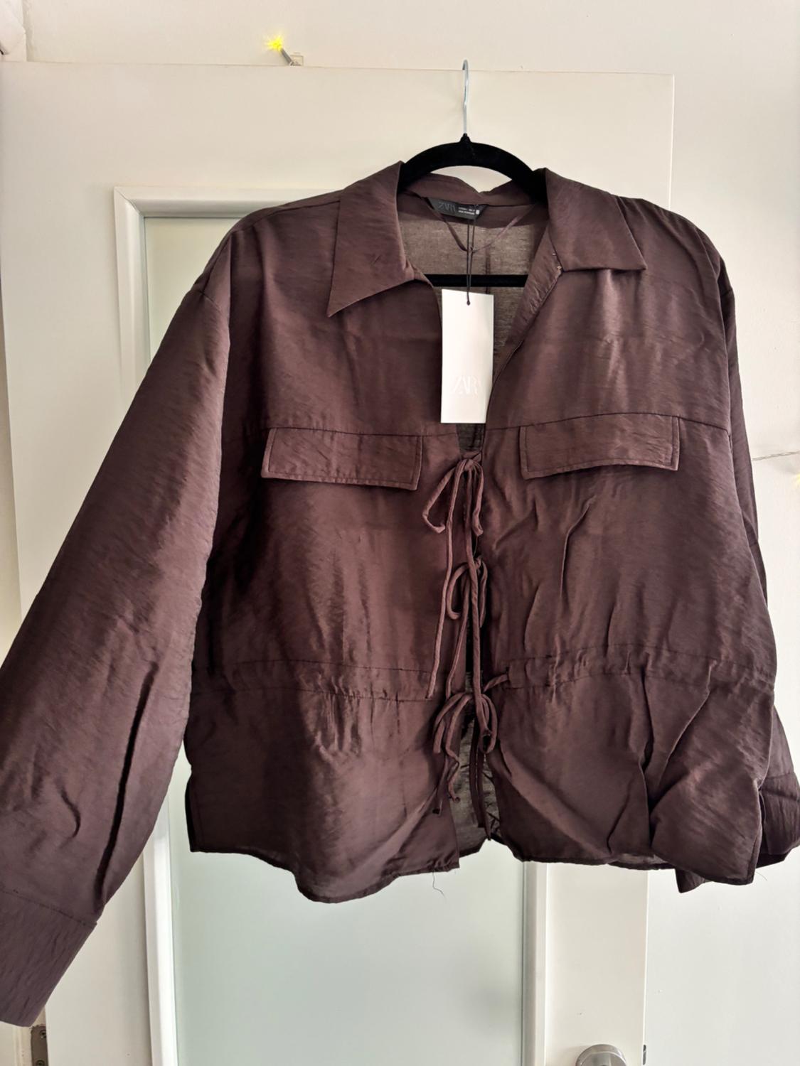 Brown jacket with lacing on the front, hanging on a black hanger over a white door. Label visible.