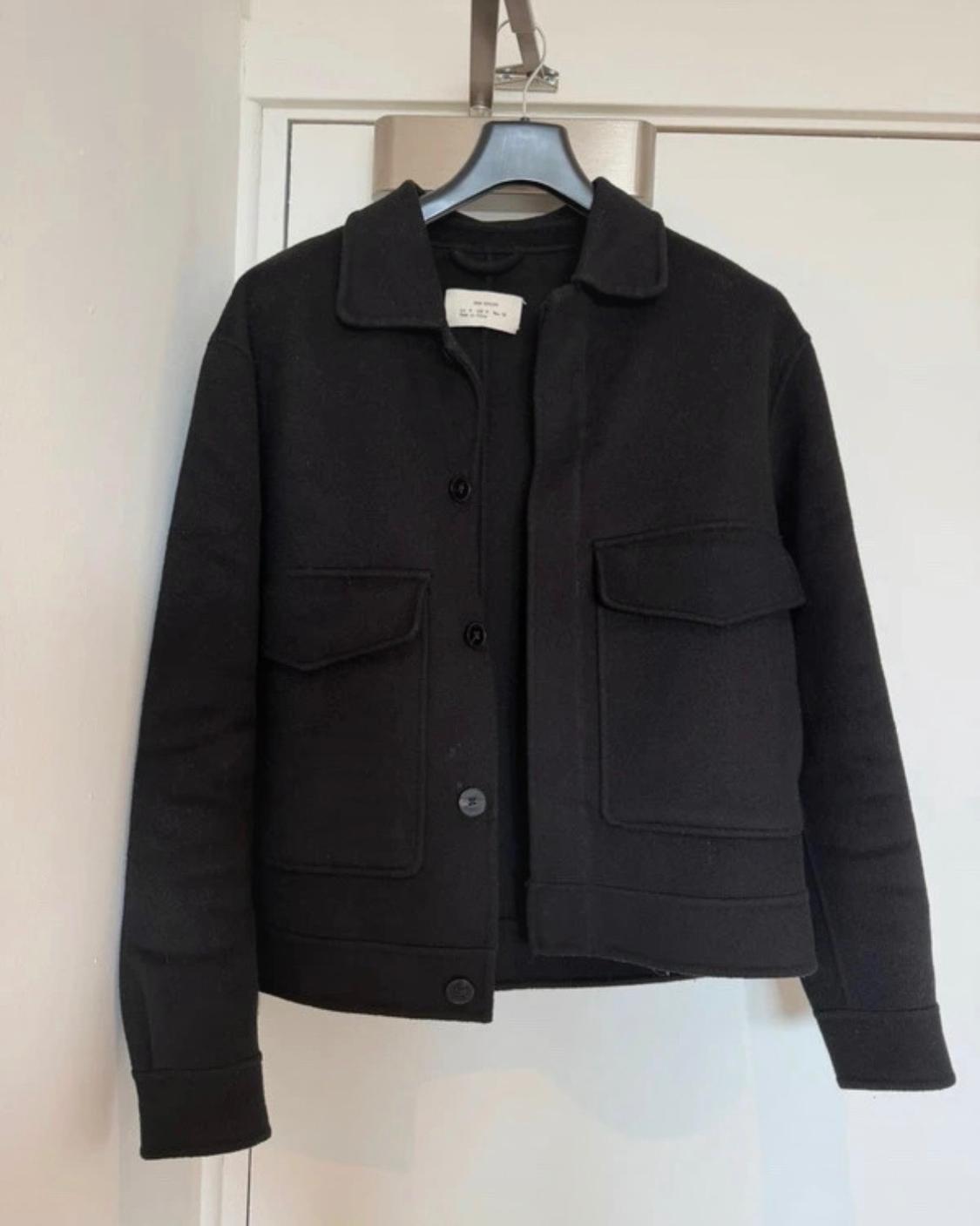 Black jacket with a collar, buttons and two large flap pockets, hanging on a black hanger on a white door.