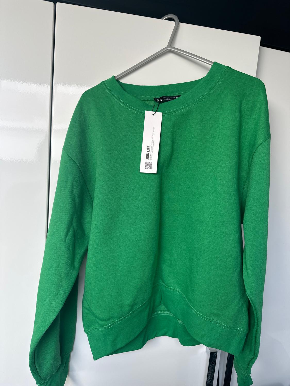 Green sweatshirt hanging on a hanger in front of a white wardrobe. Visible label with "JOIN LIFE" text and QR code.