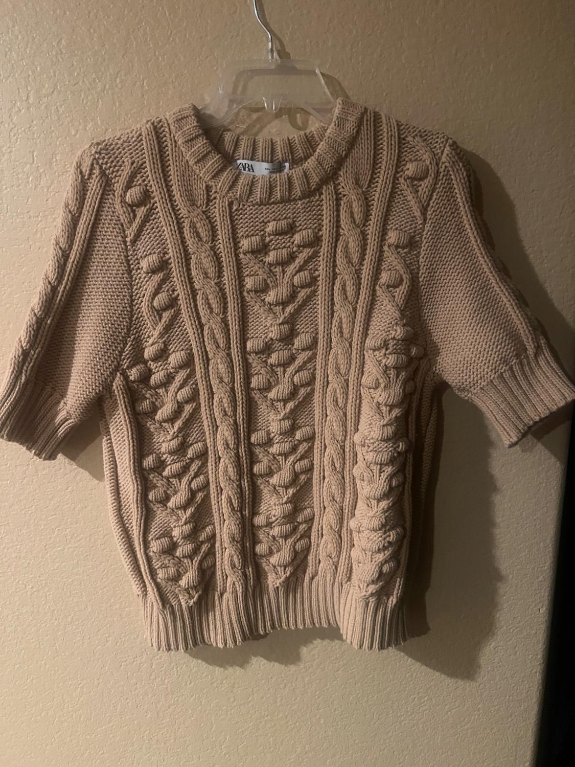 Beige knitted jumper with cable knit design and short sleeves, hanging on a clear hanger against a beige wall.
