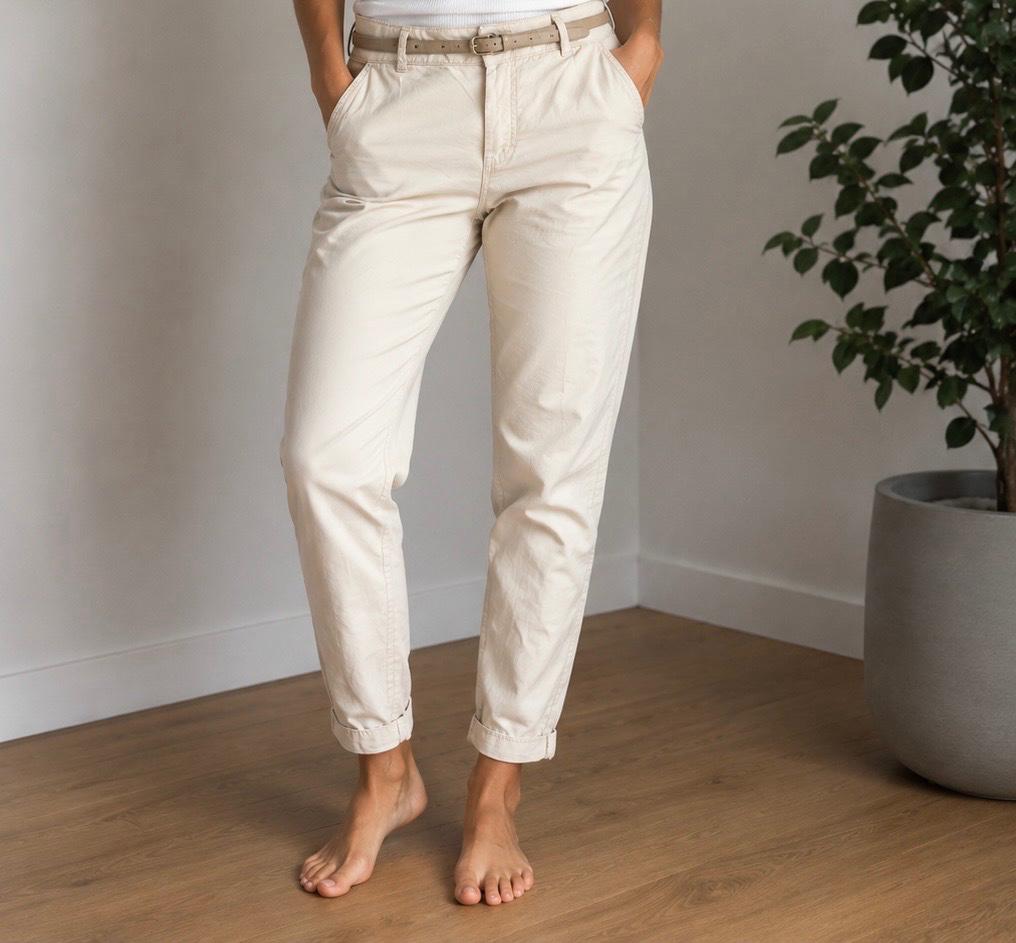 Barefoot person in beige trousers with rolled-up hems and a belt, hands in pockets. Interior with wooden flooring and a green plant.