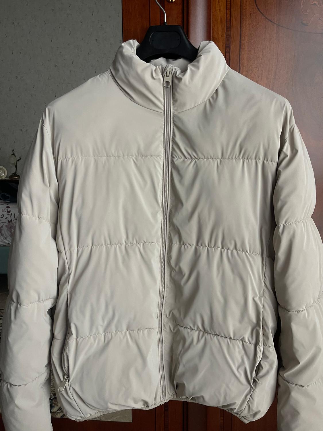 Beige padded jacket hanging on a black hanger, with a central zip, against a wooden background and grey wall.