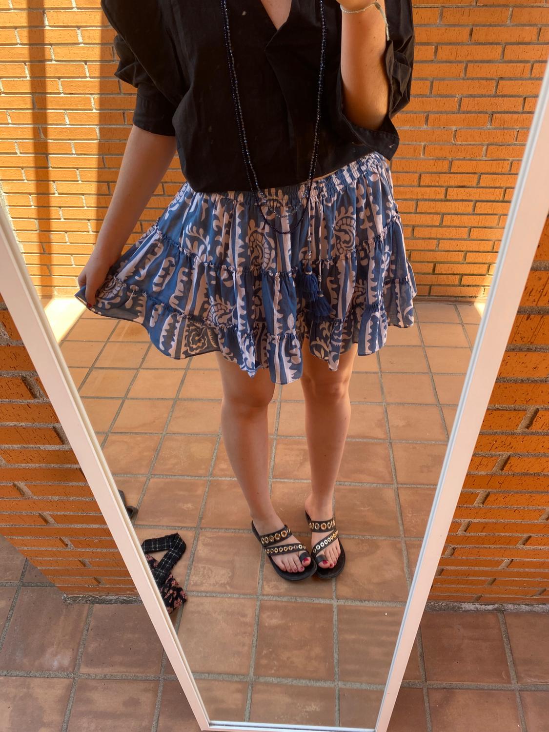 Woman wearing a black blouse and patterned blue skirt, black sandals with gold details, reflected in a mirror on a brick balcony.