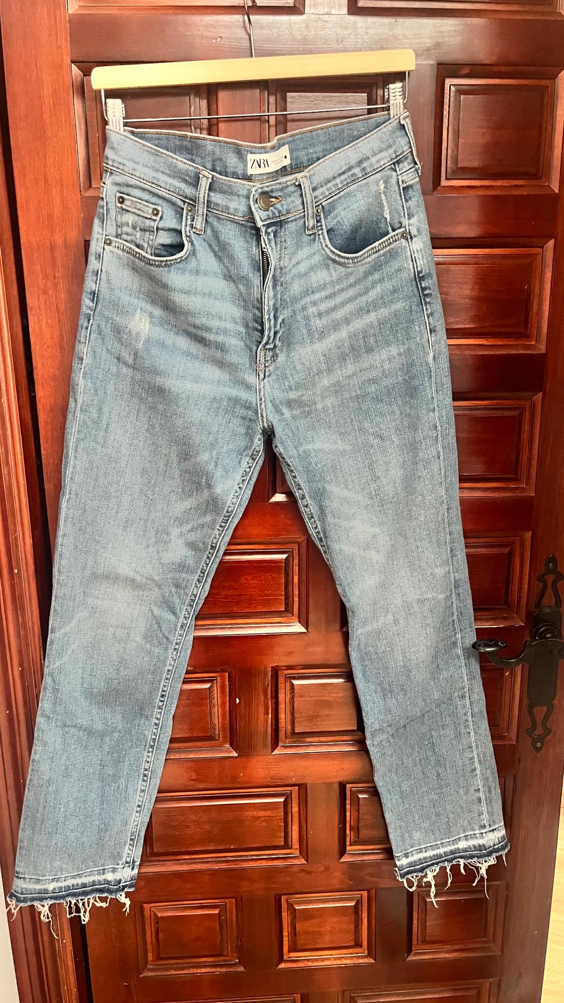 Worn blue jeans hanging on a wooden door. They have frayed hems and a visible label on the waistband.