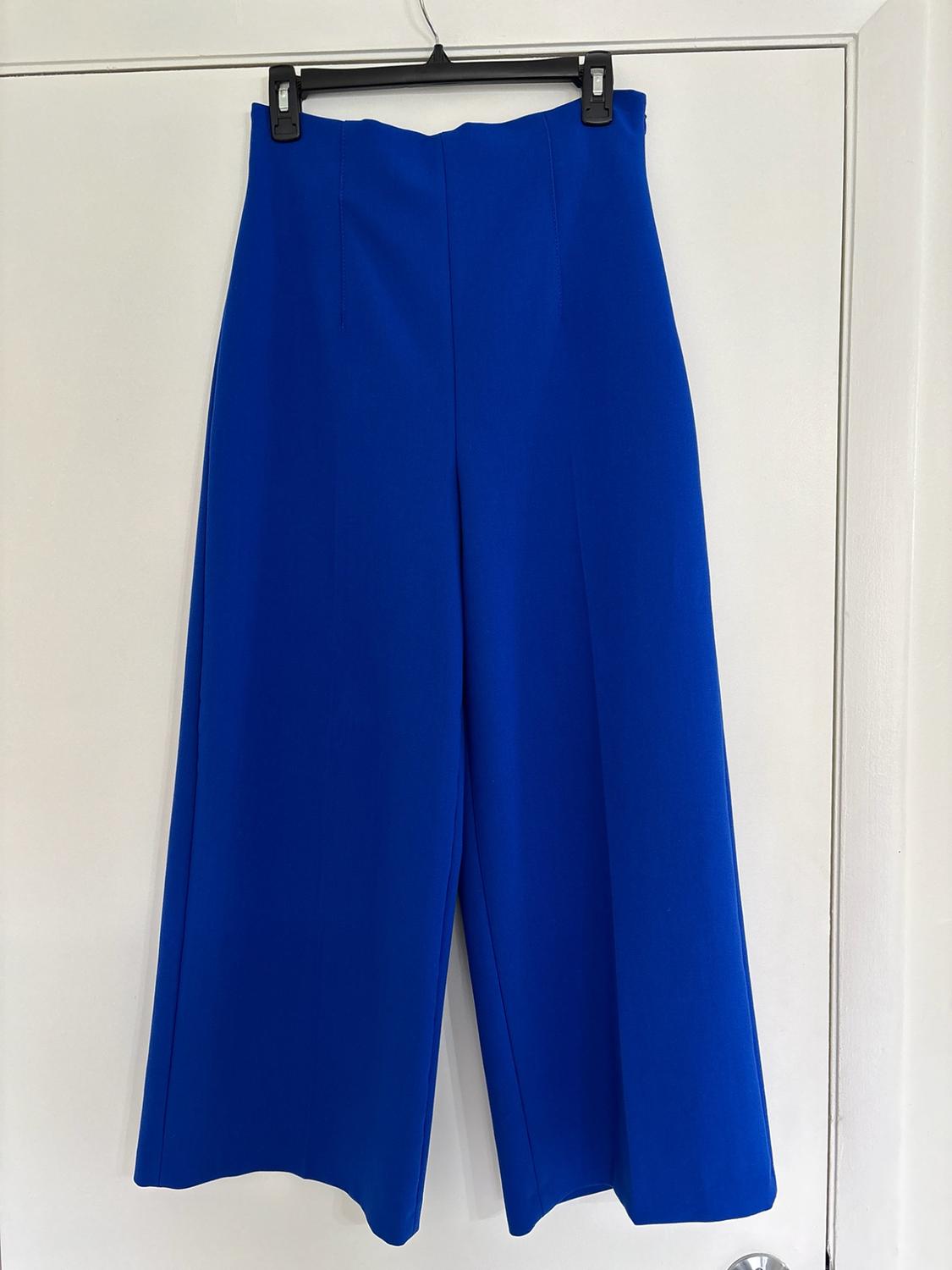High-waisted wide-leg blue trousers hanging on a black hanger in front of a white door.