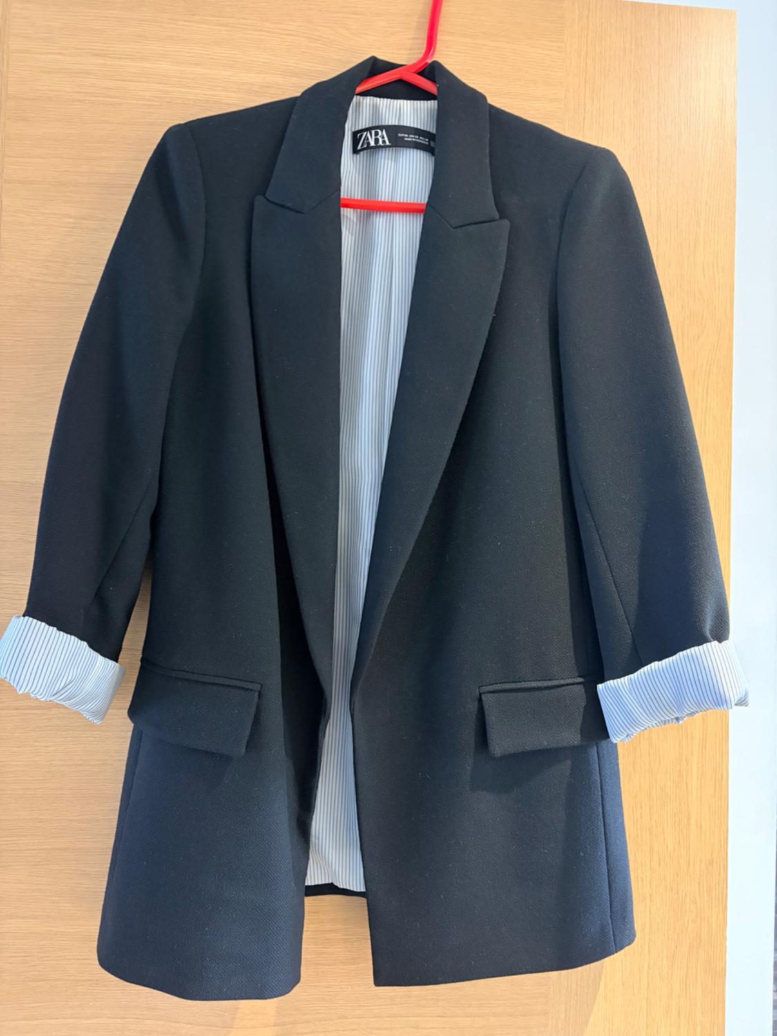 Black blazer hanging on a hanger over a wooden door. Sleeves rolled up showing striped lining. Label with the text ZARA.