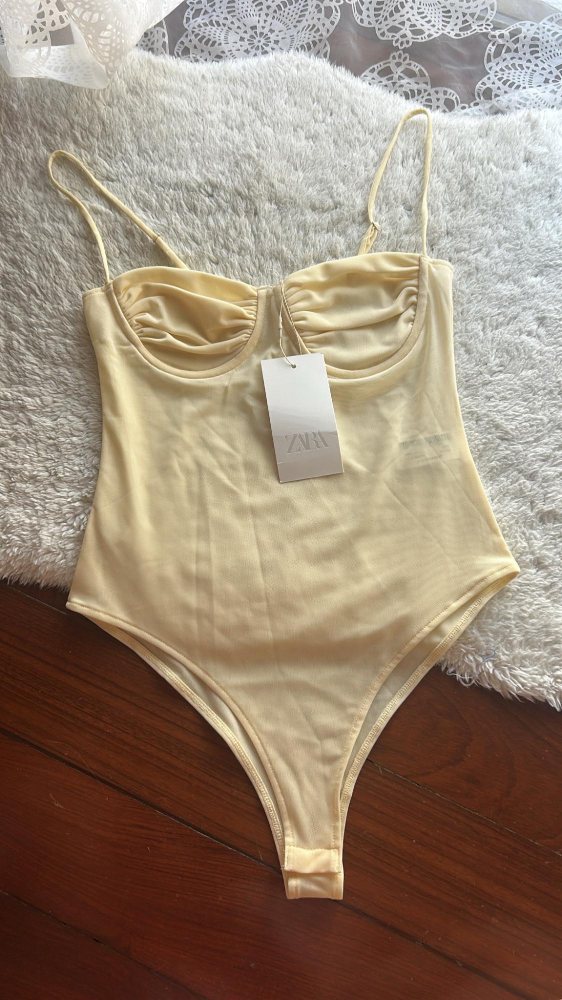 Light yellow bodysuit with thin straps and gathered cups on a white rug and wooden floor. Label with the text ZARA.