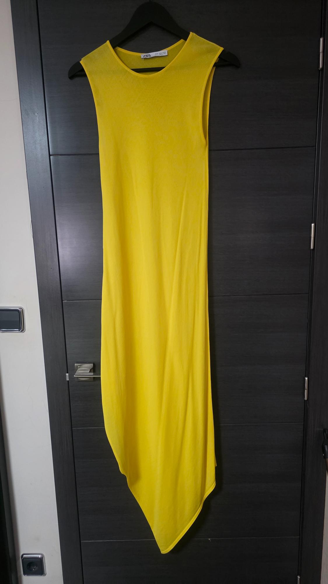 Yellow sleeveless maxi dress with an asymmetric hem hanging on a black hanger against a dark door. ZARA text on the label.