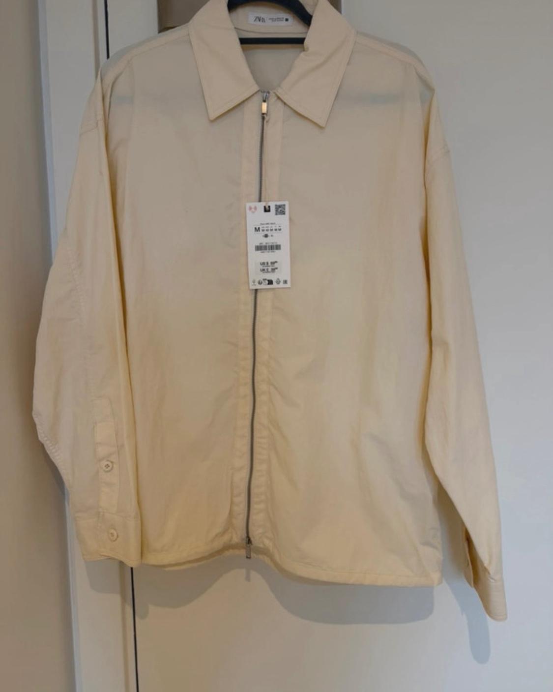 Cream-coloured lightweight jacket with a zip and collar, hanging on a hanger. Visible labels with the text ZARA and the size M.