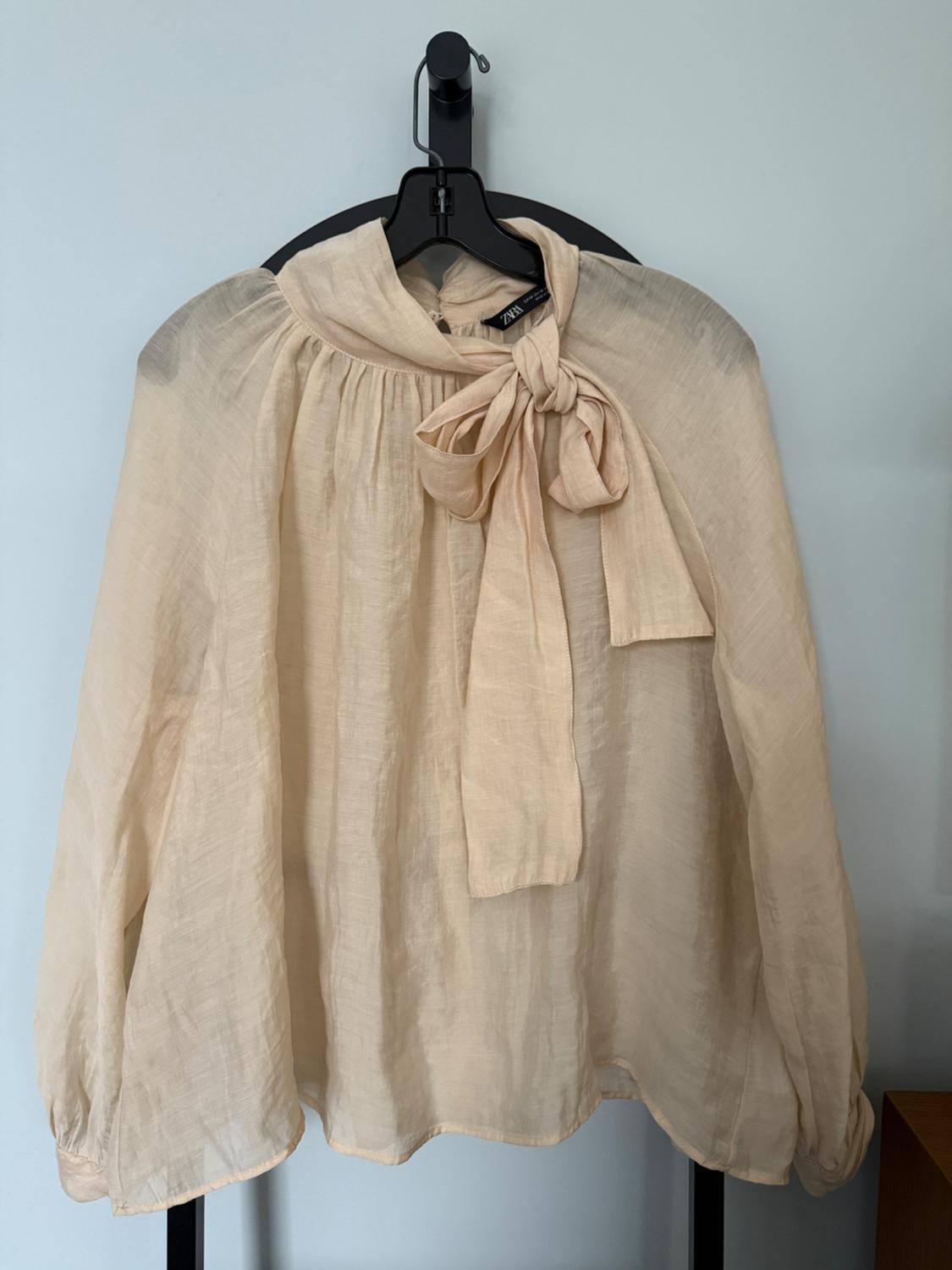 Beige lightweight fabric blouse with a bow at the neck, hanging on a black hanger. Visible label with the ZARA brand. White background.