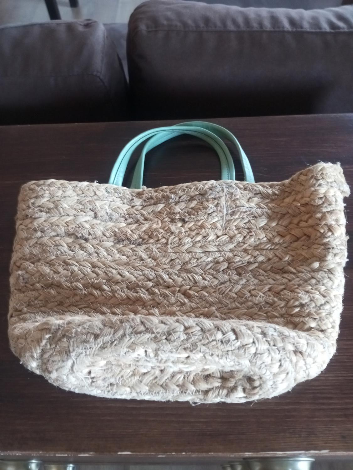 Woven straw bag with green handles on a dark wooden table, with a brown sofa in the background.
