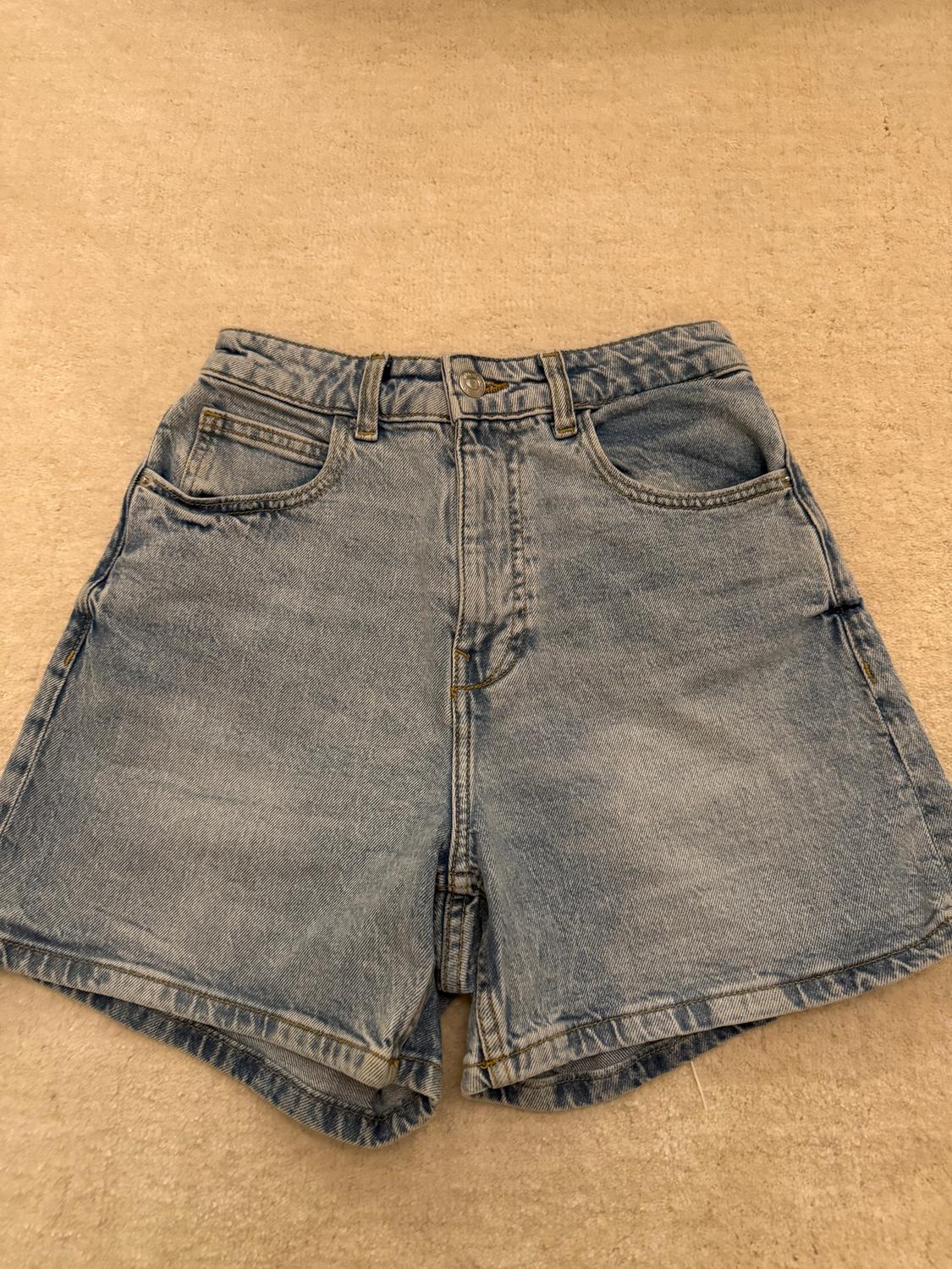 Light blue denim shorts with front pockets and belt loops, on a beige background.