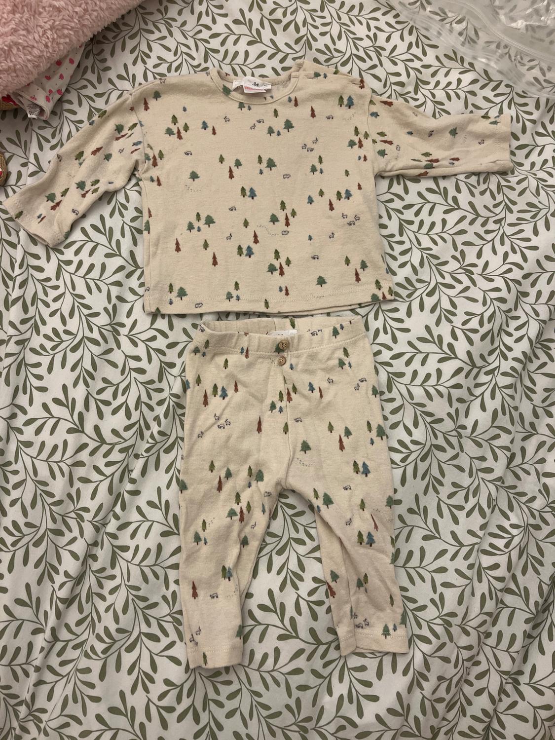 Beige children's outfit with a print of trees and small cars, on a white sheet with a green leaf design.