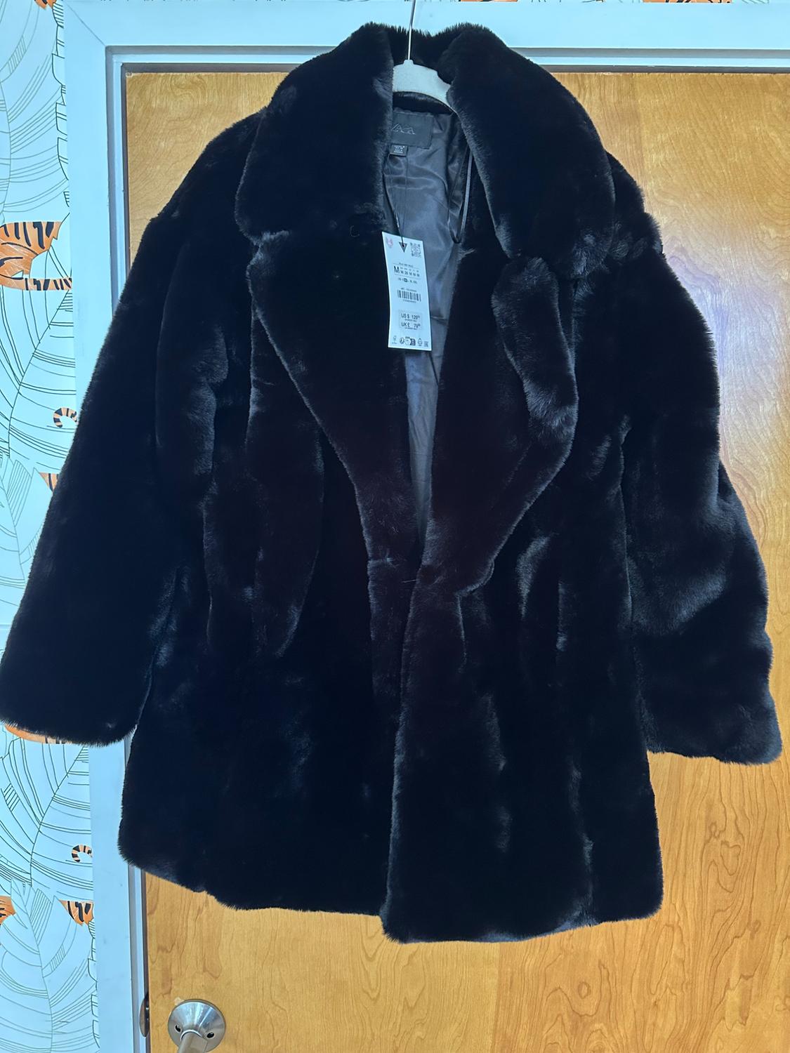 Black faux fur coat hanging on a light wooden door. Label visible inside the coat. Background with wallpaper.