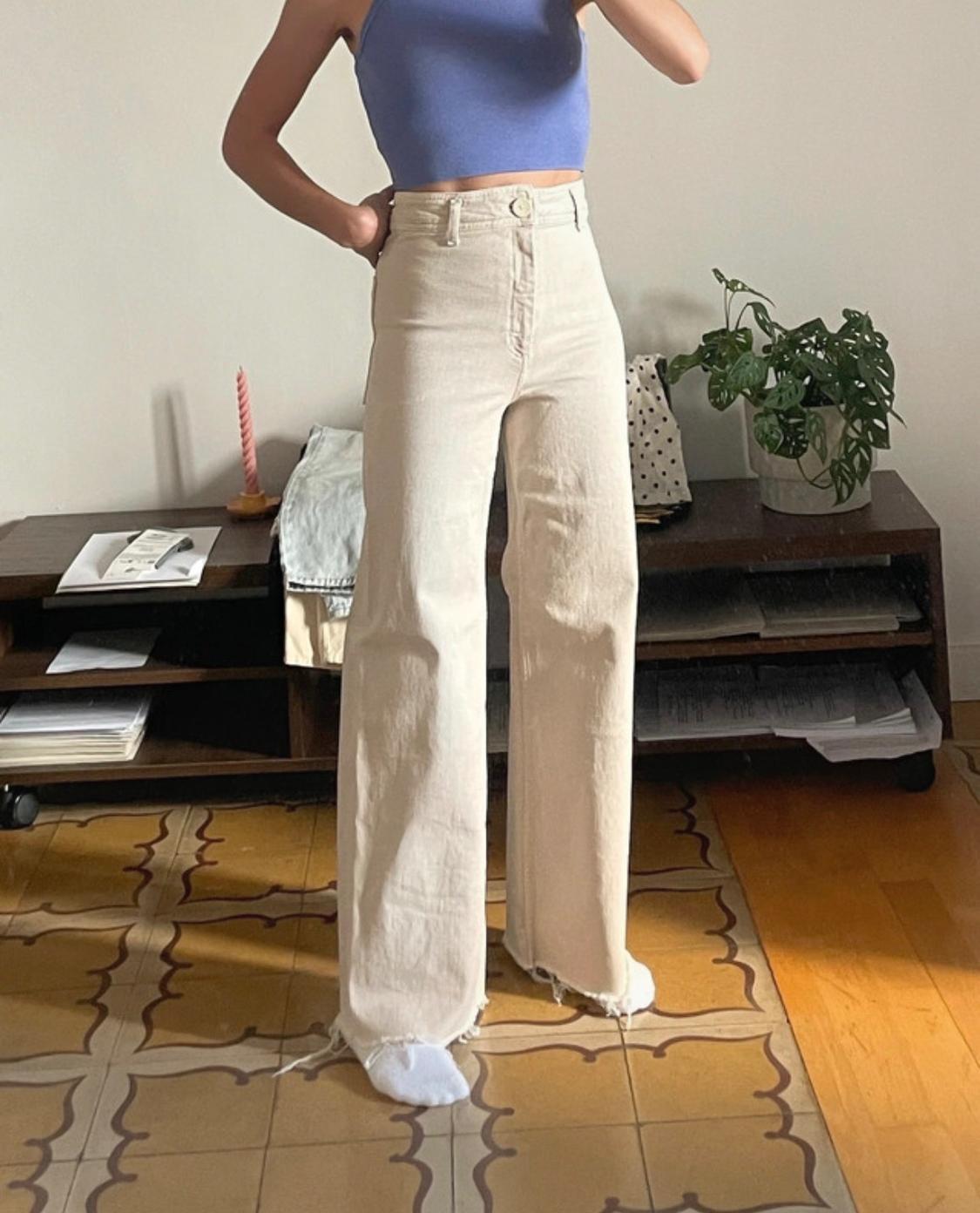 Model posing indoors wearing a blue top, high-waisted beige wide-leg jeans with a frayed hem. Background featuring a piece of furniture, a plant and a tiled floor.