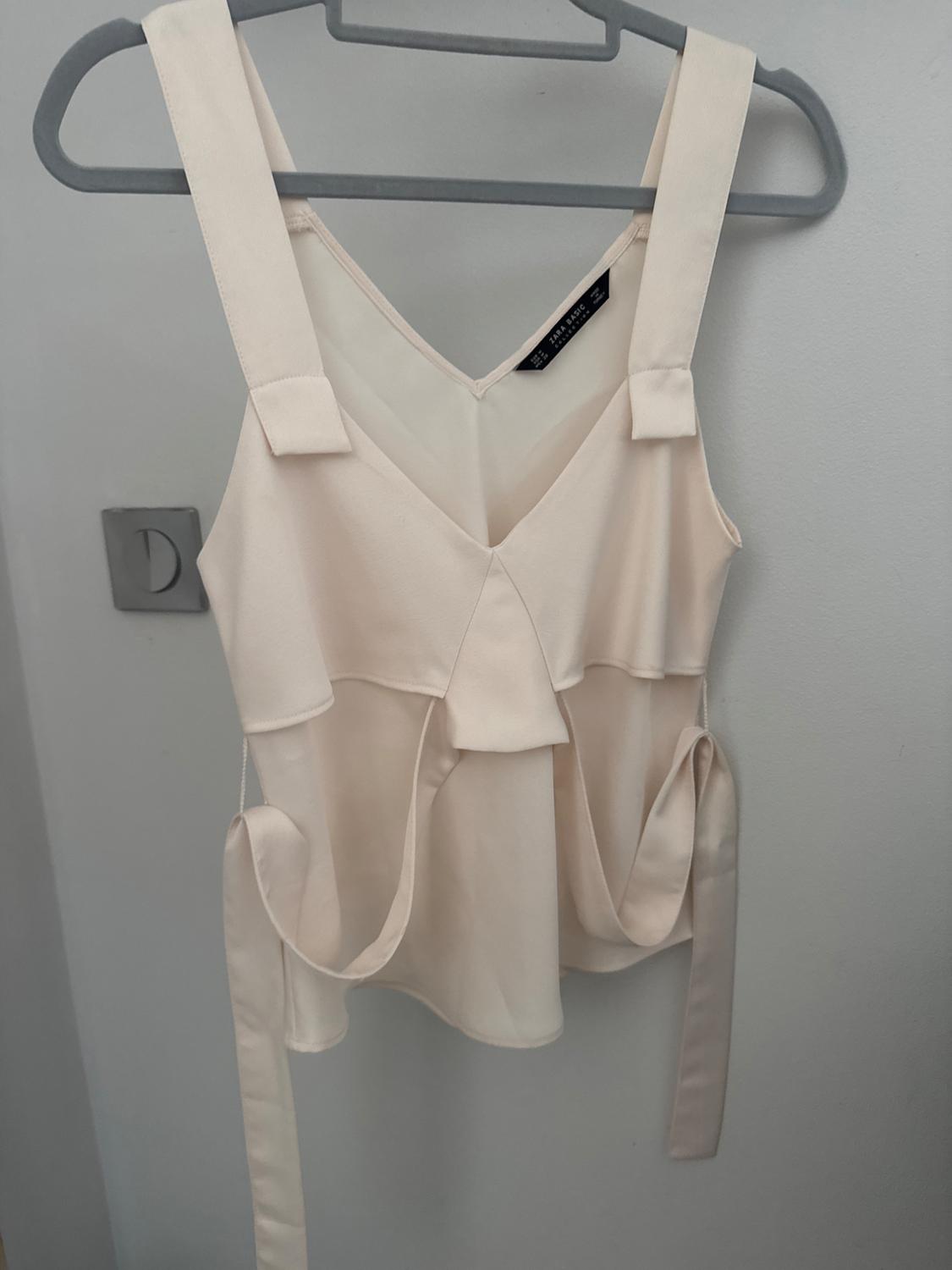 Front view of a beige blouse with wide straps and ruffle details.