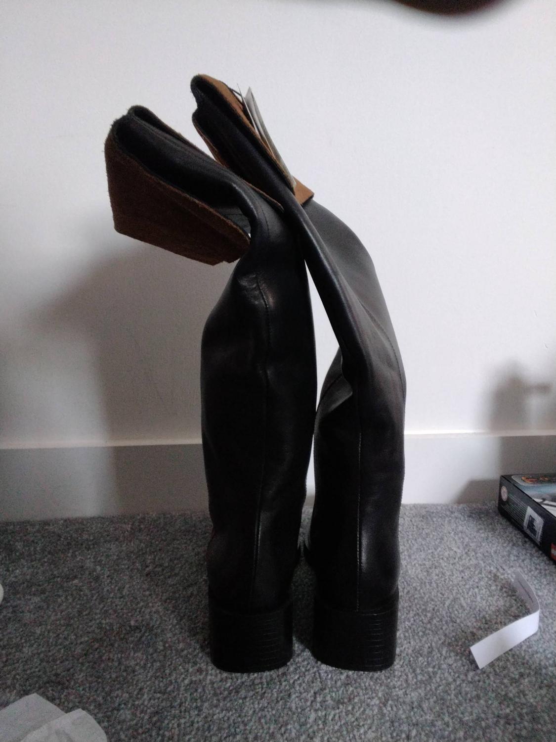 Black leather knee-high boots with the tops folded down to reveal a brown interior, standing on a grey rug next to a white wall.
