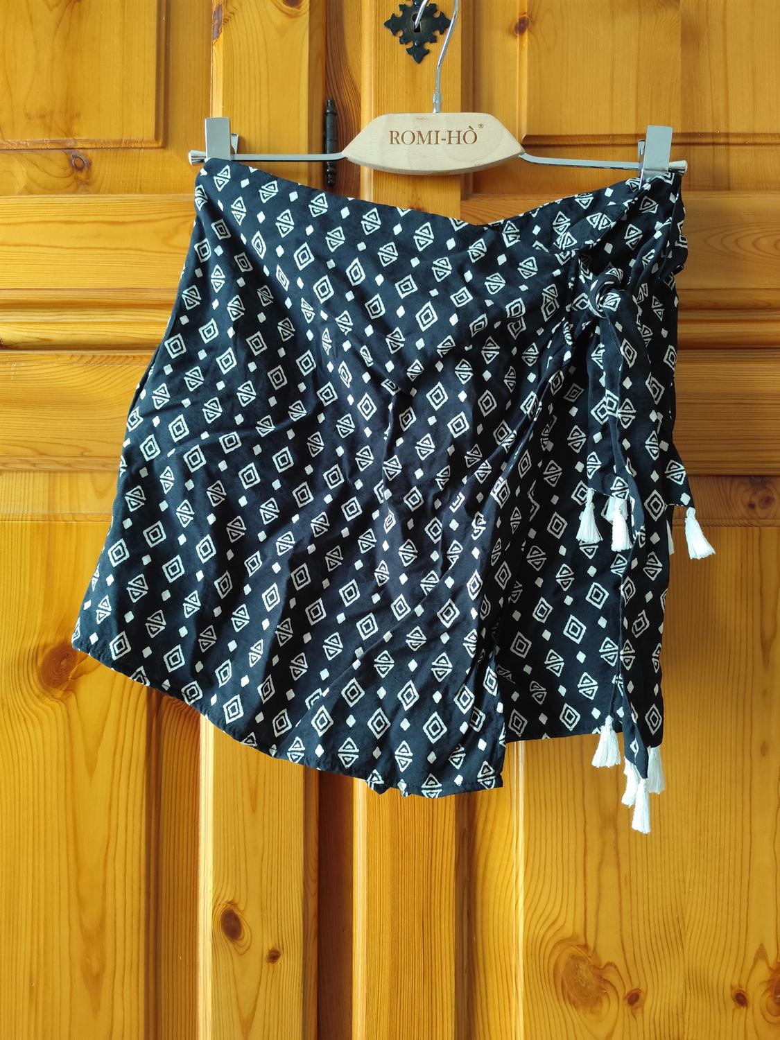 Black skirt with a white geometric print and tassels, hanging on a wooden "ROMI-HO" branded hanger against a wooden background.