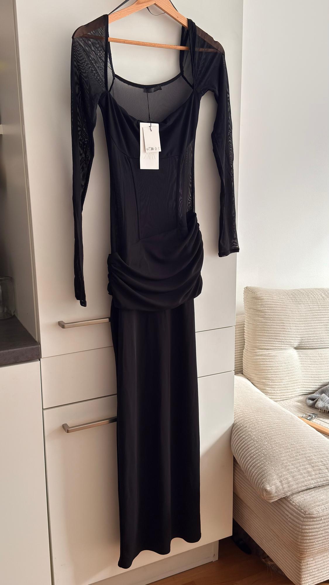 Long black dress with long mesh sleeves, hanging on a hanger over a white wardrobe. Label visible on the chest.