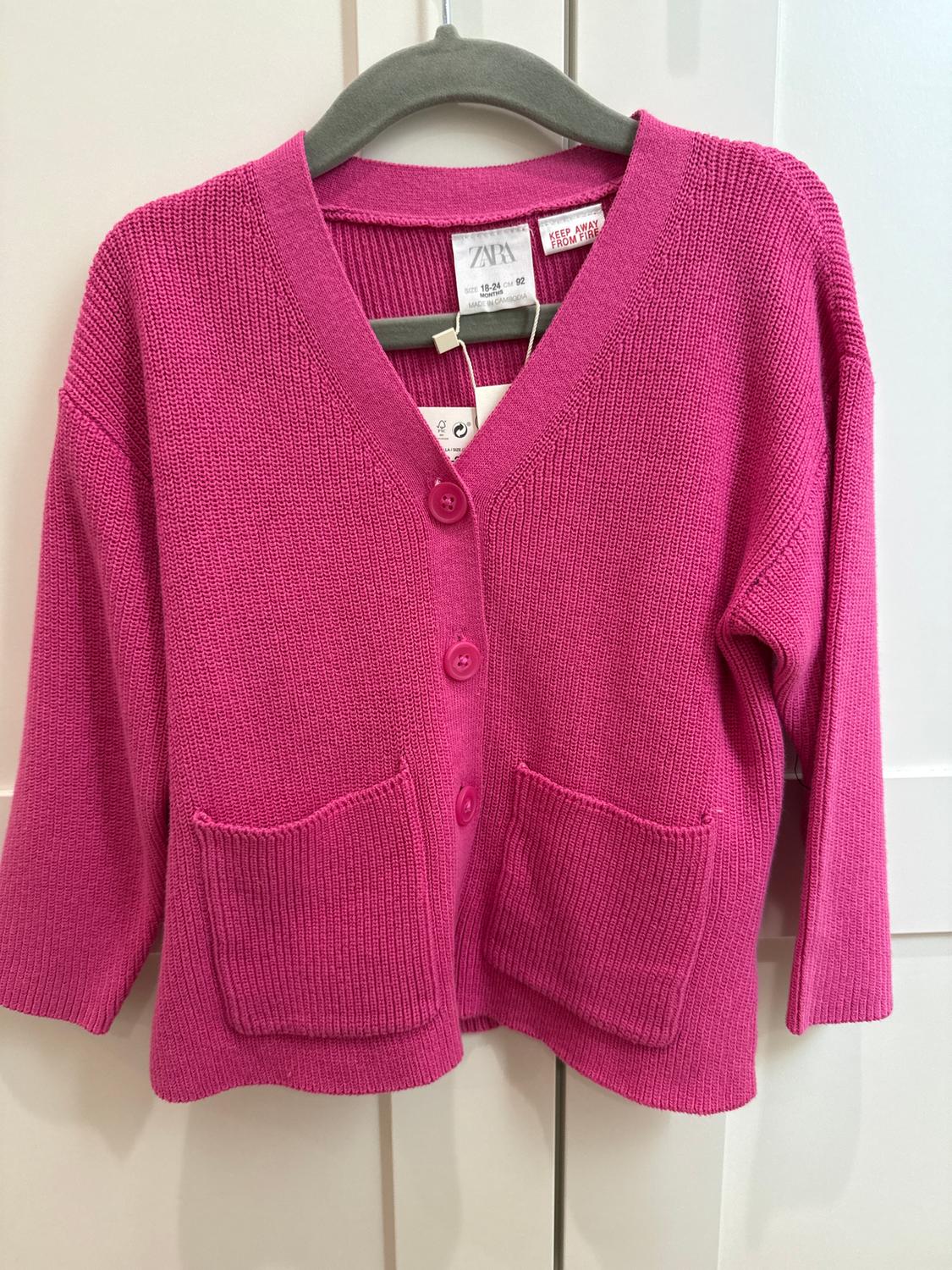 Pink knitted cardigan with buttons and front pockets, hanging on a grey hanger. Visible label: "ZARA, KEEP AWAY FROM FIRE".