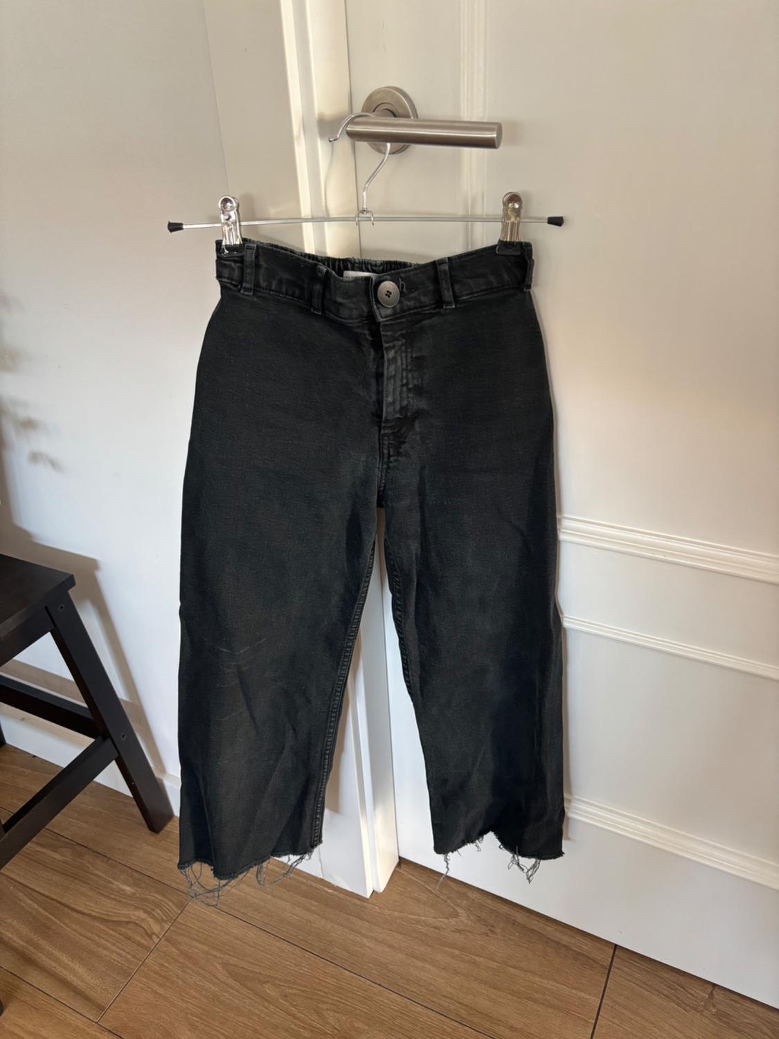 Worn black trousers hanging on a hanger over a white door, with a wooden floor and a black chair beside it.