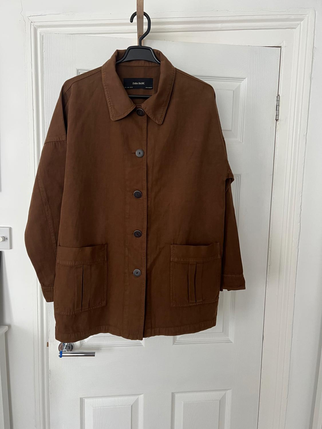 Brown jacket with buttons and front pockets, hanging on a white door. The inner label reads "ZARA BASIC".