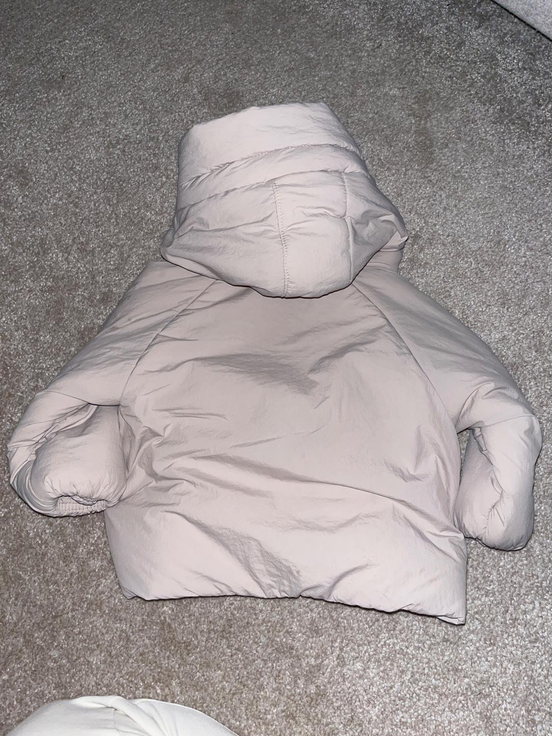 Beige padded jacket with hood on a grey rug. The garment is laid out, showcasing its voluminous and warm design.