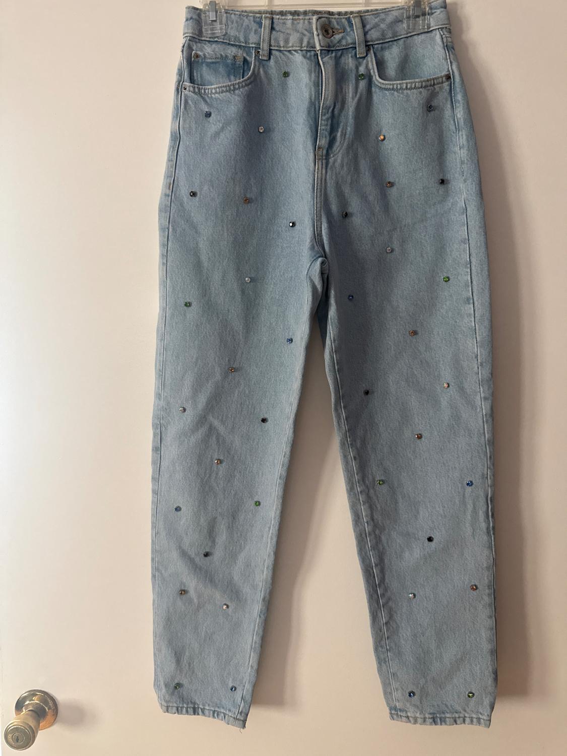 Light blue denim jeans with small, colourful sparkly details, hanging on a clear hanger over a white door.